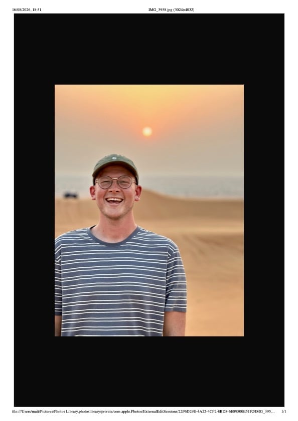 Smiling man wearing glasses and striped shirt standing in desert at sunset with sun visible in warm sky behind him